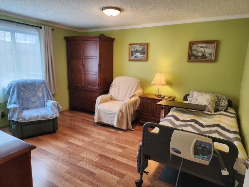 A bedroom with wood flooring, a chest of drawers with a lace doily and small plant, an open closet, a wall-mounted TV, and a wooden chest by the window—perfect for those seeking comfort or exploring senior care FAQs Snohomish County.